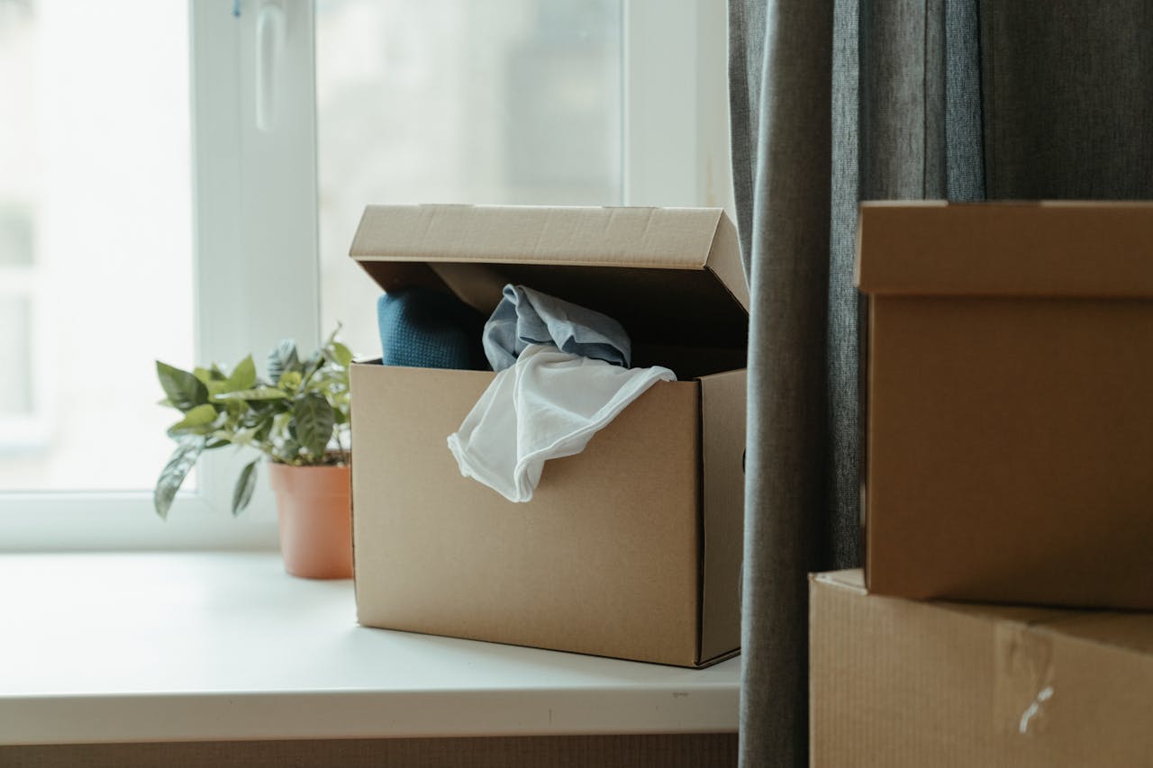 Open cardboard box with clothes by window, symbolizing moving or relocation.